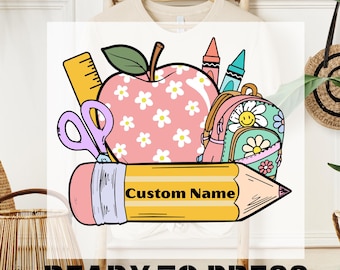 May include: A DTF transfer design featuring school-themed graphics. The design includes a pink apple with white flowers, a backpack, scissors, a ruler, crayons, and a pencil with the text "Custom Name". The words "DTF TRANSFER" and "READY TO PRESS" are also included.