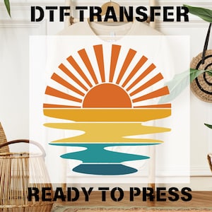 May include: A white t-shirt with a DTF transfer design featuring a sunset graphic. The design includes an orange sun with radiating lines, and layers of yellow, teal, and blue representing the ocean. The text "READY TO PRESS" is at the bottom.