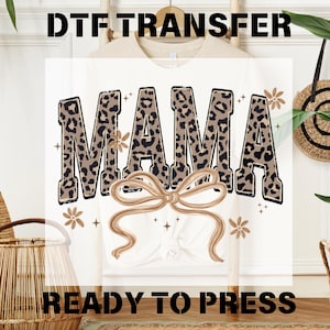 May include: White t-shirt featuring the word "MAMA" in a leopard print design, with a bow and floral accents. The text "DTF TRANSFER" and "READY TO PRESS" are displayed above and below the main design.