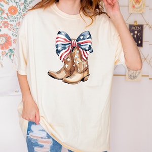 May include: Cream-colored t-shirt featuring a graphic of brown cowboy boots adorned with white stars. A large bow, patterned with red and white stripes and blue stars, sits atop the boots. The shirt is a casual, oversized fit.