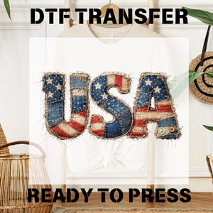 May include: White t-shirt with the word "USA" in a patchwork design featuring the American flag. The letters are made of denim and fabric with red, white, and blue patterns. The text "DTF TRANSFER READY TO PRESS" is above and below the design.