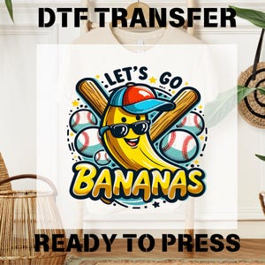 May include: White t-shirt with a cartoon banana wearing a baseball cap and sunglasses, holding two baseball bats. The banana is surrounded by baseballs and the text "Let's Go Bananas".