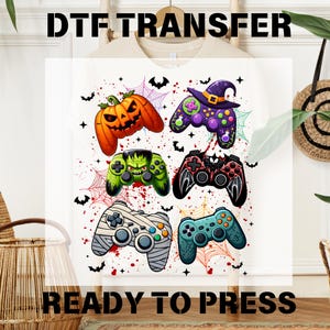 May include: White t-shirt with a DTF transfer design featuring six Halloween-themed video game controllers. The controllers are designed as a jack-o'-lantern, witch, Frankenstein, mummy, and more. The text "DTF TRANSFER" and "READY TO PRESS" are displayed.