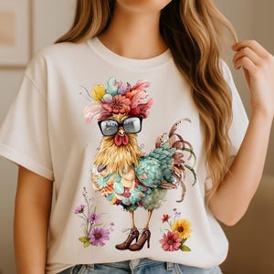 May include: Cream-colored t-shirt featuring a whimsical rooster illustration. The rooster wears glasses and a floral headdress, with colorful feathers and a patterned body. The design includes flowers and high-heeled shoes, creating a unique and artistic look.