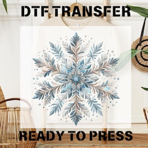 May include: A white t-shirt with a blue and brown snowflake design. The design is intricate and detailed, with a glittery finish. The shirt is ready to be pressed with a heat press.