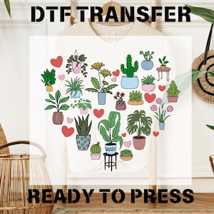 May include: A white t-shirt with a heart-shaped design featuring a variety of potted plants and flowers. The text "DTF TRANSFER" appears at the top of the image and "READY TO PRESS" appears at the bottom.