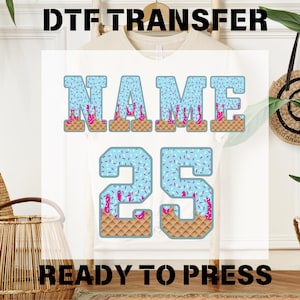 May include: White t-shirt with a DTF transfer design featuring the word "NAME" and the number "25" in a blue, pink, and brown ice cream cone design. The text "READY TO PRESS" is printed at the bottom.