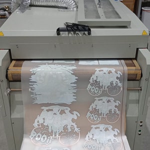 May include: A large industrial printer with a roll of printed material. The material features white graphics of Halloween-themed designs, including a spider web and the word "BOO!" The printer is gray and black.