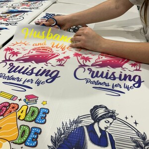 May include: A person is cutting a yellow vinyl decal that says "Husband and Wife" above a cruise ship graphic. The image also shows other decals with text and graphics, including "Cruising Partners for life" and a school-themed design.