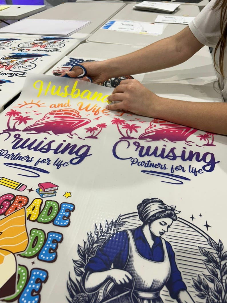 May include: A person is cutting a yellow vinyl decal that says "Husband and Wife" above a cruise ship graphic. The image also shows other decals with text and graphics, including "Cruising Partners for life" and a school-themed design.