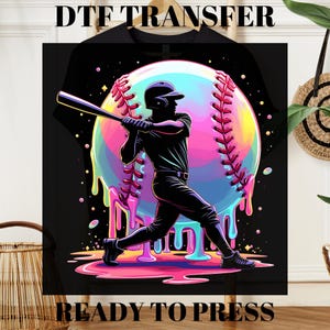 May include: Black baseball-themed graphic with a batter swinging a bat, set against a colorful baseball. The baseball has a rainbow-colored design with dripping paint effects. The text "DTF TRANSFER" and "READY TO PRESS" are at the top and bottom.
