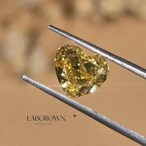 May include: A heart-shaped yellow lab-grown diamond held by tweezers. The diamond is clear and has a slight yellow hue. The text "LABGROWN GERMANY" is visible in the image.