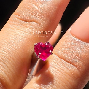 Peut inclure: Une bague en argent avec une pierre précieuse en rubis de laboratoire de forme cœur. La bague est sur un doigt.