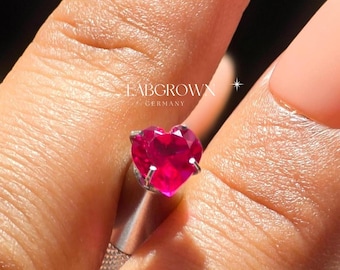 Puede incluir: Un anillo de plata con una piedra preciosa de rubí de laboratorio en forma de corazón. El anillo está en un dedo.