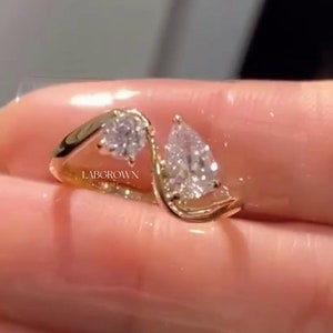 Könnte beinhalten: Ein goldener Ring mit zwei birnenförmigen Diamanten. Der Ring hat ein gebogenes Band und die Diamanten sind in einer Krappenfassung gefasst. Der Ring ist mit "LABGROWN" gekennzeichnet.