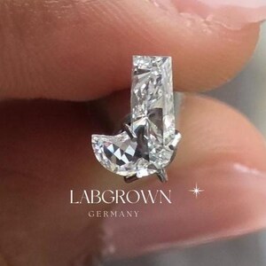 Puede incluir: Un diamante claro cultivado en laboratorio con forma de la letra "J" se sostiene entre dedos con esmalte de u&ntilde;as rosa. El diamante est&aacute; facetado y refleja la luz. El texto "LABGROWN GERMANY" es visible.