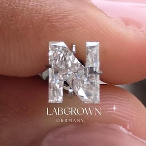 Pode incluir: Um close-up de um anel de diamante com uma pedra central de corte retangular e duas pedras laterais menores. O anel é cravejado em uma banda de prata. O anel está rotulado como "LABGROWN GERMANY".