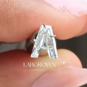 Puede incluir: Se muestra un diamante de laboratorio transparente con forma de la letra "A". El diamante est&aacute; facetado y es rectangular. El texto "LABGROWN GERMANY" es visible debajo. El fondo est&aacute; borroso.