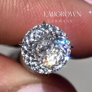Op de afbeelding: Een close-up van een ronde, heldere diamanten ring gezet in een zilveren band. De ring zit op een vinger met roze nagellak. De tekst "LABGROWN GERMANY" is zichtbaar in de afbeelding.