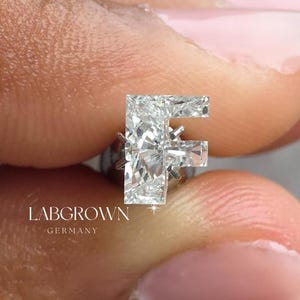 Może przedstawiać: Wisiorek z diamentem wyhodowanym w laboratorium w kształcie litery "F". Wisiorek trzymany jest między palcami na szarym tle. Wisiorek wykonany jest z przezroczystych, błyszczących diamentów. Widoczny jest tekst "LABGROWN GERMANY".