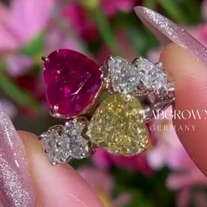 May include: A close-up of a ring with three heart-shaped gemstones. The ring features a large red heart-shaped gemstone, a smaller yellow heart-shaped gemstone, and a cluster of smaller white heart-shaped gemstones. The ring is set in white metal and has the text "Labgrown Germany" on the band.