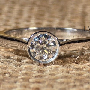 Peut inclure: Une bague simple en argent avec un diamant de laboratoire rond brillant serti clos. La bague est sur un fond en toile de jute brune. LABGROWN DIAMONDS est imprimé sur le côté droit de l'image.