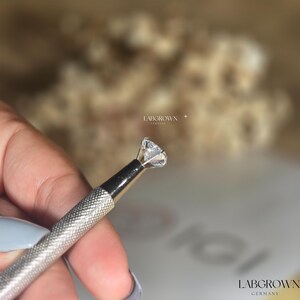 May include: A close-up of a clear, round diamond held in a metal tool. The diamond is being examined for quality and clarity. The text "LABGROWN GERMANY" is visible in the background.