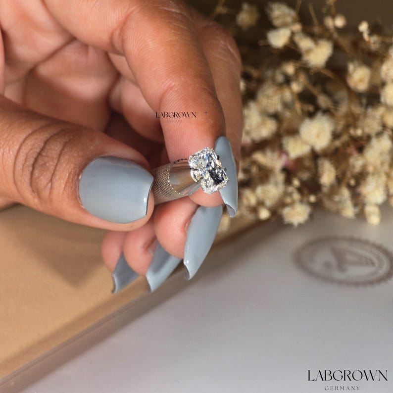 May include: A close-up of a clear, square-shaped diamond gemstone set in a silver ring setting. The ring is held by a hand with light-colored nail polish. The text "LABGROWN" is visible in the background.