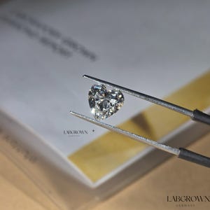 May include: A heart-shaped lab-grown diamond held by tweezers. The diamond is clear and sparkly. LABGROWN GERMANY is printed on the box in the background.