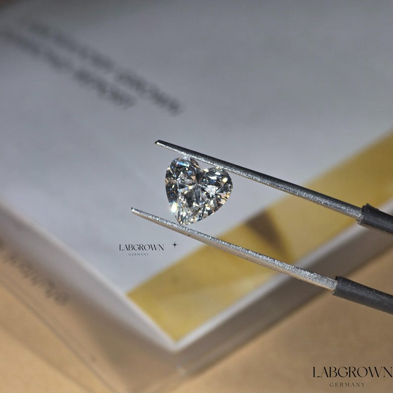 May include: A heart-shaped lab-grown diamond held by tweezers. The diamond is clear and sparkly. LABGROWN GERMANY is printed on the box in the background.