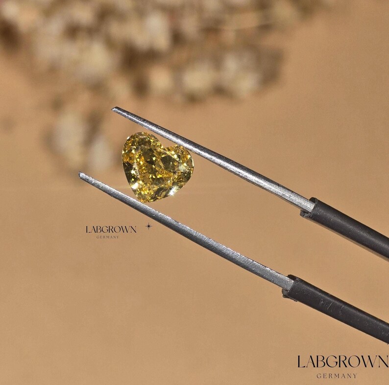 May include: A heart-shaped yellow lab-grown diamond held by tweezers. The diamond is clear and has a slight yellow hue. The text "LABGROWN GERMANY" is visible in the image.