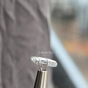 May include: A clear, lab-grown diamond held by a silver tool. The diamond is rectangular with multiple facets, reflecting light. The text "LABGROWN GERMANY" is visible in the background.