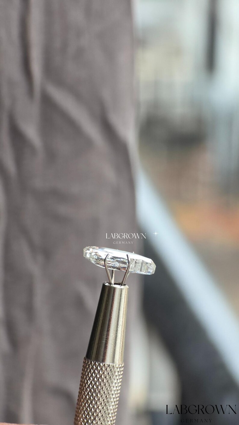 May include: A clear, lab-grown diamond held by a silver tool. The diamond is rectangular with multiple facets, reflecting light. The text "LABGROWN GERMANY" is visible in the background.