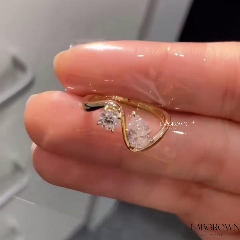 May include: A gold ring with two clear gemstones. The ring is designed with a wavy band and the gemstones are set in a prong setting. The ring is on a finger.