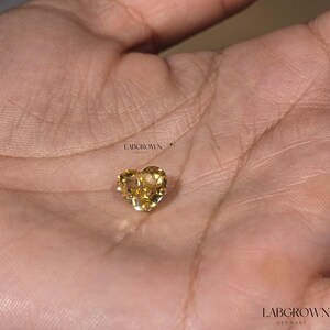 May include: A heart-shaped, yellow lab-grown diamond, set in a silver setting. The diamond is displayed on a hand.