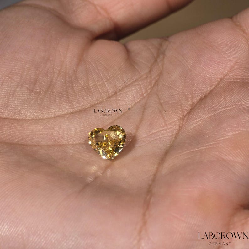 May include: A heart-shaped, yellow lab-grown diamond, set in a silver setting. The diamond is displayed on a hand.