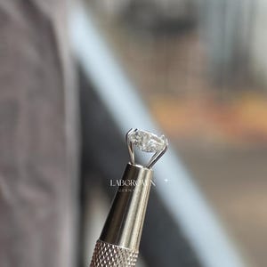 May include: An oval-cut lab-grown diamond ring held in a silver tool. The diamond is set in a silver band. The tool has a textured handle. The text "LABGROWN GERMANY" is visible.