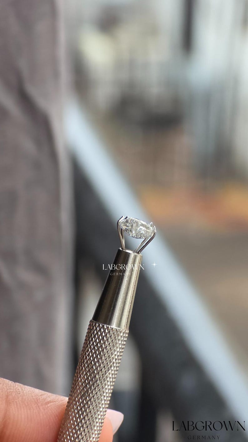 May include: An oval-cut lab-grown diamond ring held in a silver tool. The diamond is set in a silver band. The tool has a textured handle. The text "LABGROWN GERMANY" is visible.
