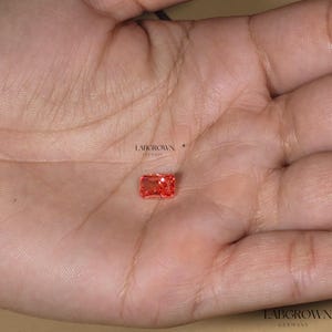 Puede incluir: Una gema rectangular, de color rojo anaranjado, cultivada en laboratorio, se sostiene en una mano. La gema tiene un corte radiante y se coloca sobre un fondo neutro. El texto "LABGROWN GERMANY" es visible.