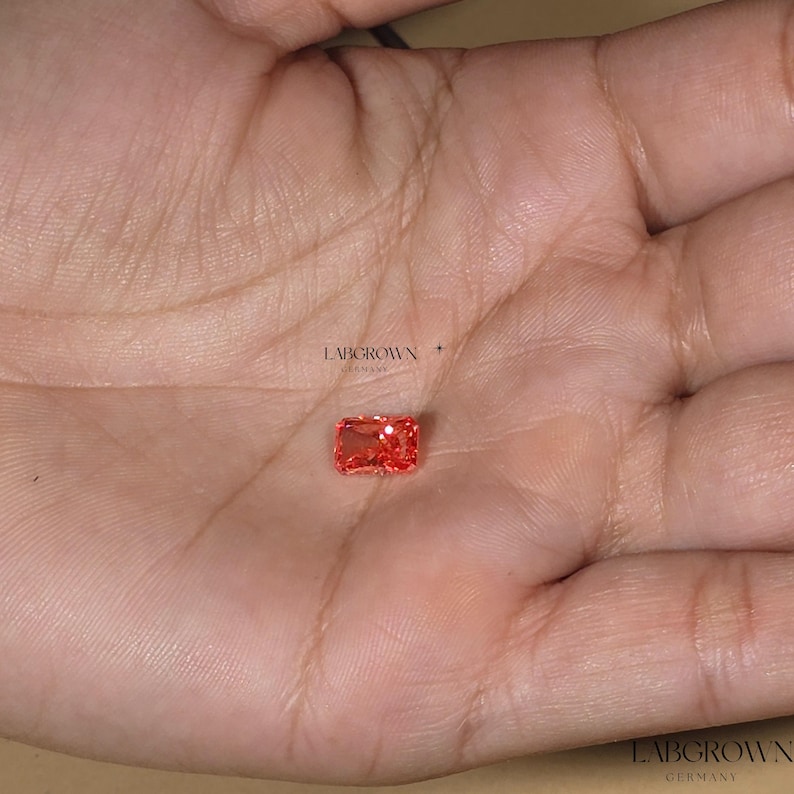 Puede incluir: Una gema rectangular, de color rojo anaranjado, cultivada en laboratorio, se sostiene en una mano. La gema tiene un corte radiante y se coloca sobre un fondo neutro. El texto "LABGROWN GERMANY" es visible.