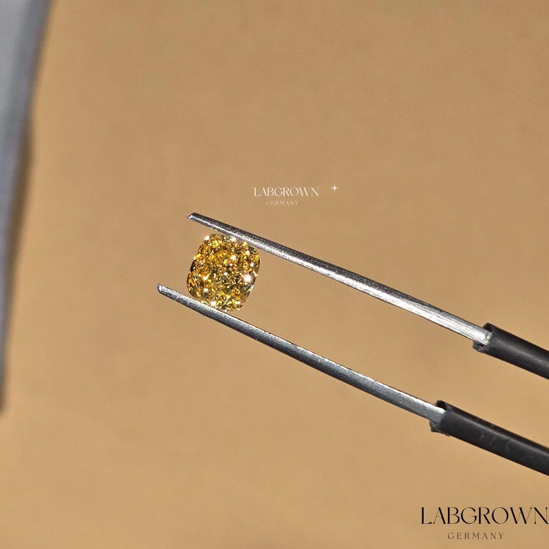 May include: A yellow lab-grown diamond held by tweezers. The diamond is a cushion cut and has a bright, golden color.