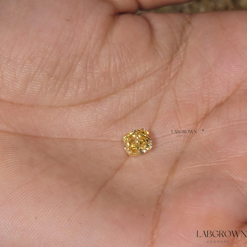 May include: A yellow cushion-cut lab-grown diamond on a person's hand. LABGROWN GERMANY is printed on the hand.
