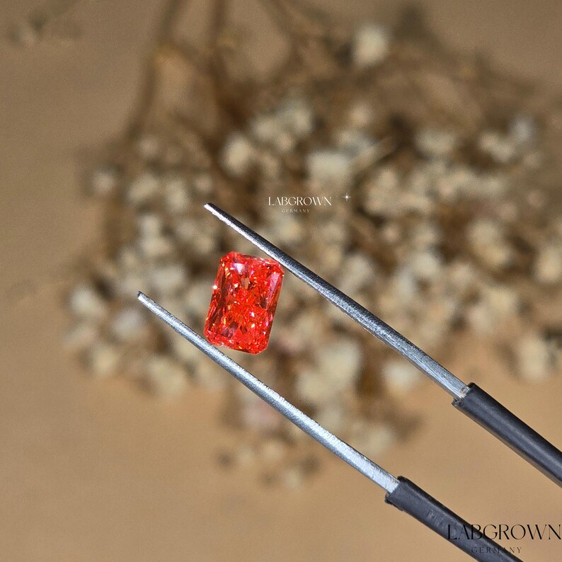 Puede incluir: Un vibrante diamante rojo cultivado en laboratorio, de forma rectangular, es sujetado por unas pinzas. La gema tiene una superficie brillante y texturizada. El fondo es una suave mezcla borrosa de beige y blanco, con el texto "LABGROWN GERMANY".