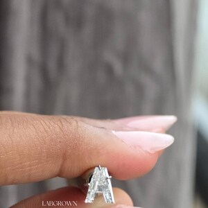 Puede incluir: Un pendiente de diamante cultivado en laboratorio con forma de la letra "A". El pendiente est&aacute; engastado en un metal plateado. El texto "LABGROWN GERMANY" est&aacute; impreso en el pendiente. El fondo es gris borroso.