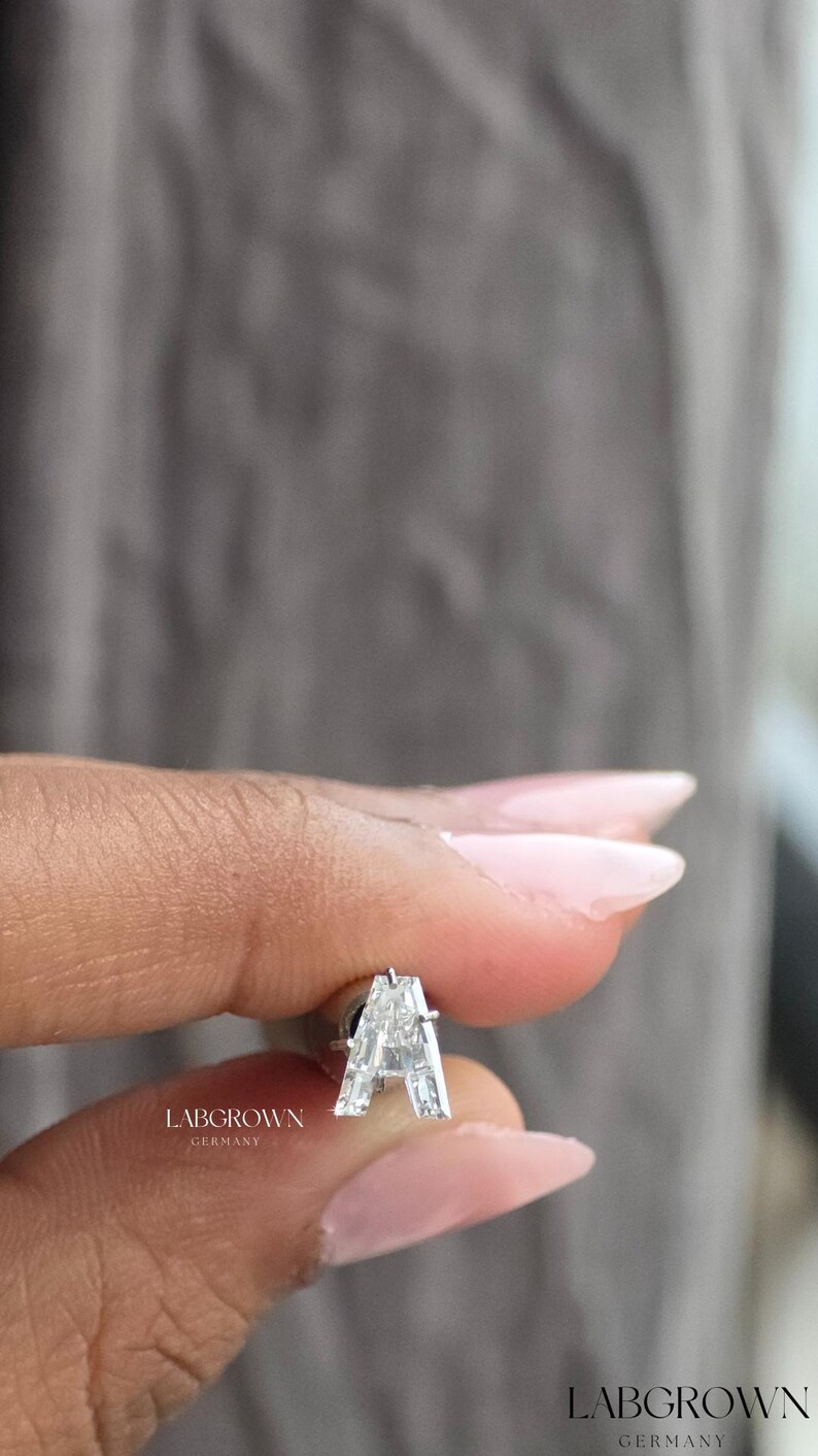 Puede incluir: Un pendiente de diamante cultivado en laboratorio con forma de la letra "A". El pendiente est&aacute; engastado en un metal plateado. El texto "LABGROWN GERMANY" est&aacute; impreso en el pendiente. El fondo es gris borroso.