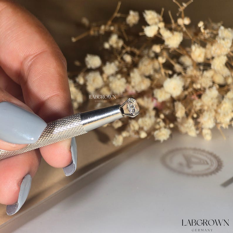 May include: A close-up of a clear, round, lab-grown diamond held in a silver tool. The tool has the text "LABGROWN GERMANY" printed on it.