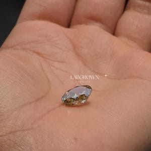 以下が含まれることがあります： 明るい茶色の色合いの、マーキス型のラボグロウンダイヤモンドが、開いた手のひらに乗っています。 ダイヤモンドには複数のファセットがあり、光を反射しています。 テキスト「LABGROWN GERMANY」が表示されています。