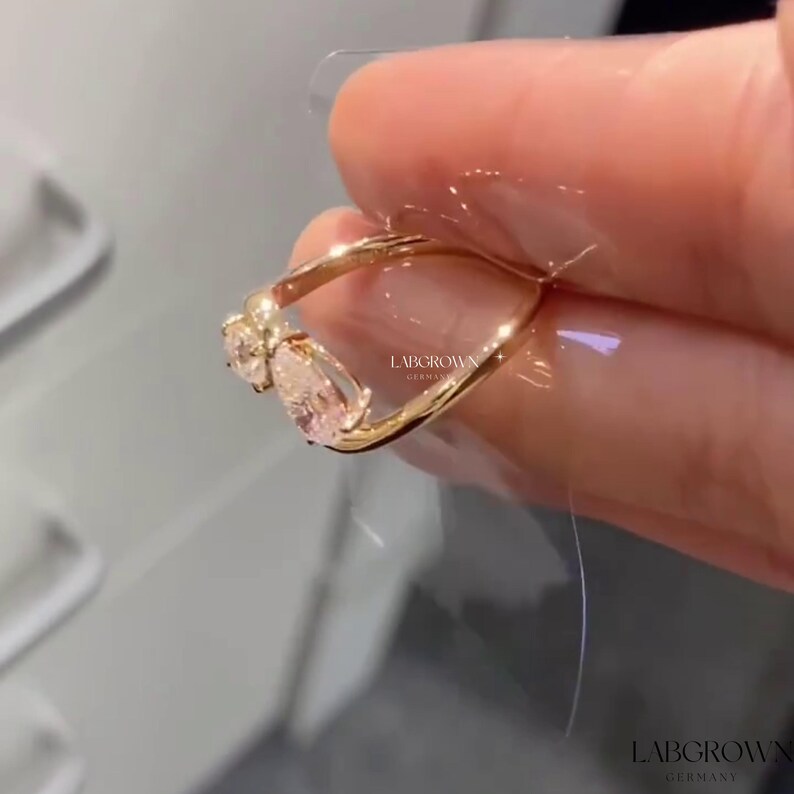 May include: A gold ring with two pear-shaped clear stones set in a twisted band. The ring is on a finger and the brand name "LABGROWN GERMANY" is visible.