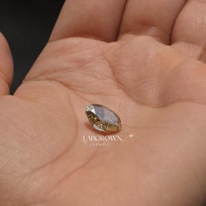 以下が含まれることがあります： 手の中に収まる、シャンパンカラーのラボグロウンオーバルカットダイヤモンド。ダイヤモンドにはファセットが見え、わずかに輝いています。「LABGROWN GERMANY」の文字が下に表示されています。