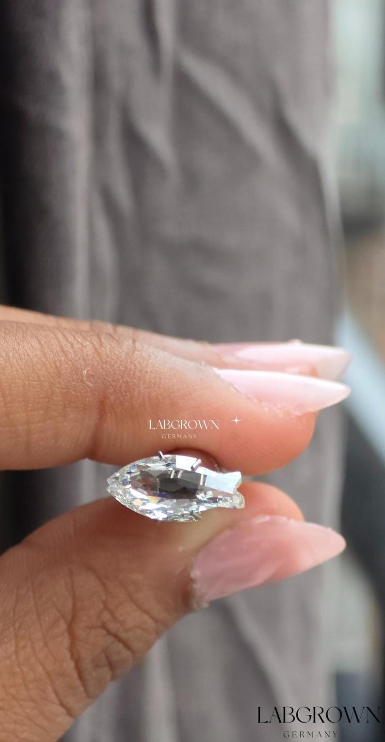 May include: A clear, pear-shaped gemstone with multiple facets, held between fingers. The gemstone reflects light, creating a sparkling effect. The text "LABGROWN GERMANY" is visible.